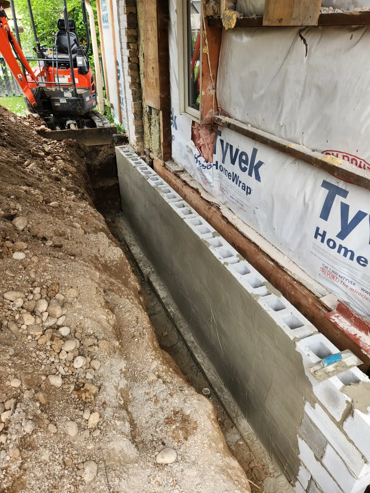 Block foundation wall exposed and coated during exterior excavation, with a mini excavator on site