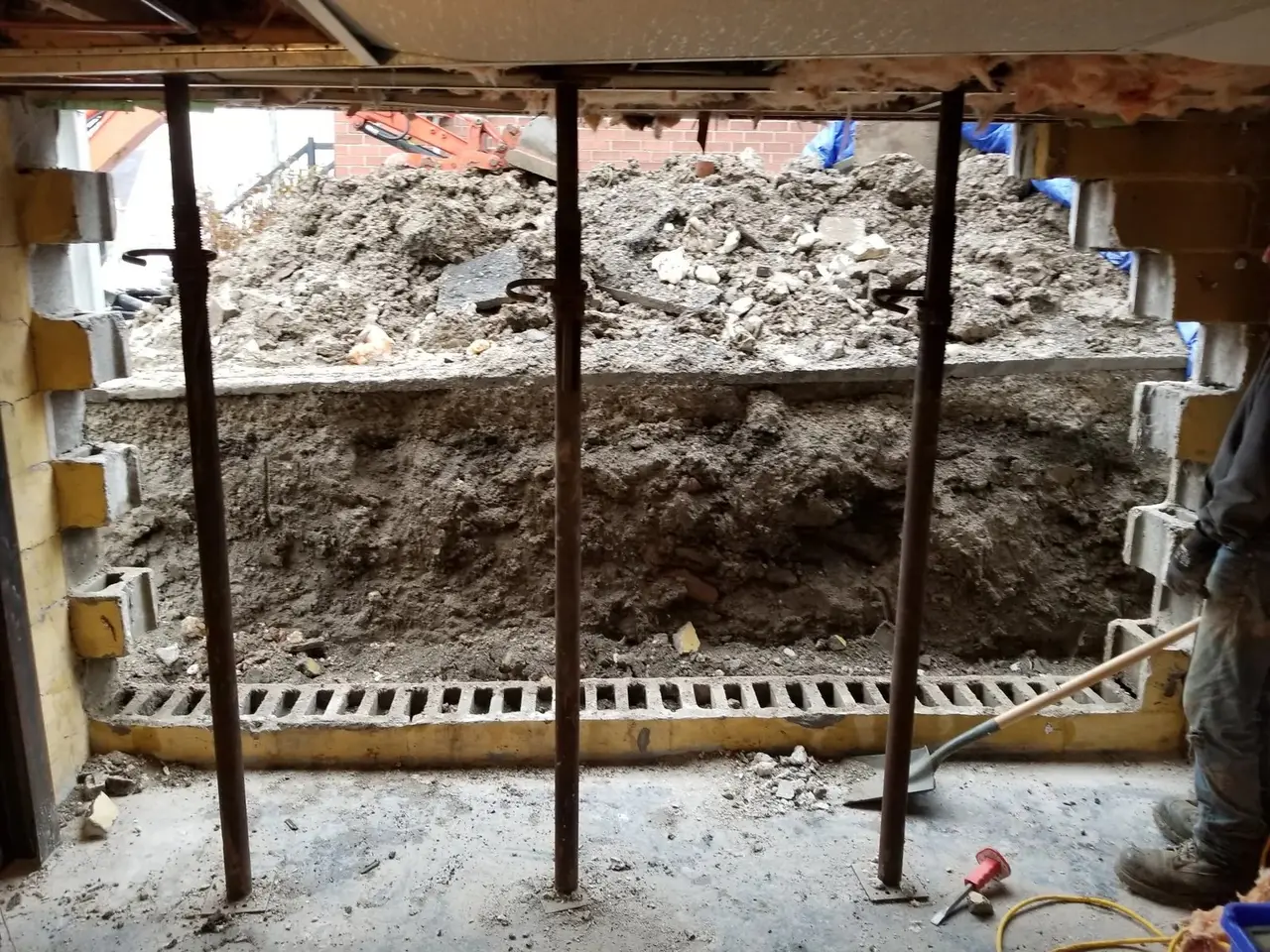 Basement underpinning in progress: soil bench excavated under a block foundation wall with temporary steel shoring
